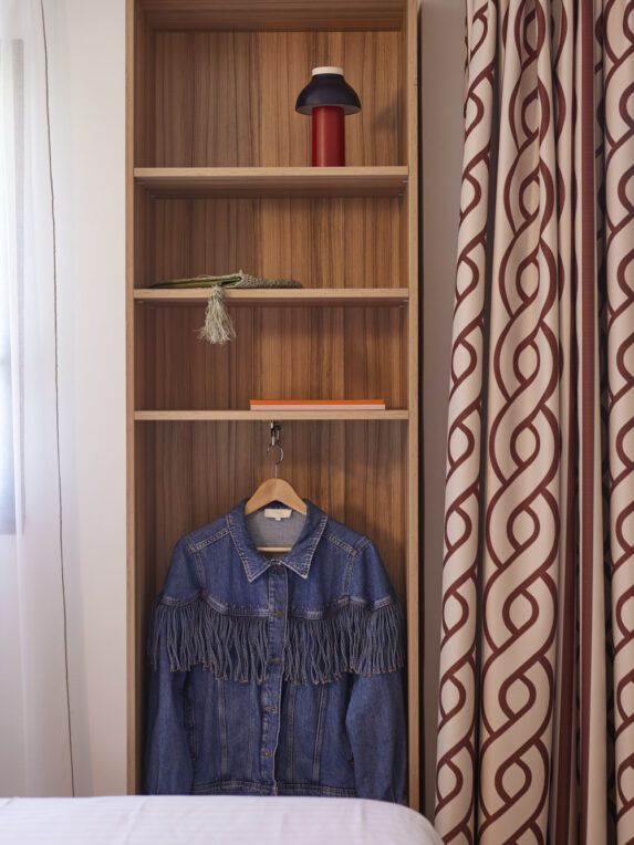 Étagère en bois ouverte avec lampe et objets décoratifs, veste en jean suspendue et rideaux à motifs sur le côté.