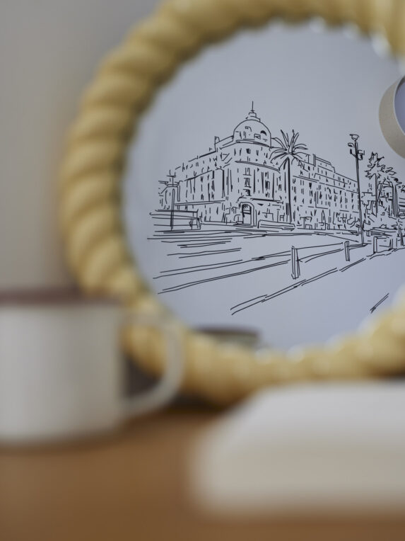 Miroir rond à bord tressé reflétant un dessin mural d’architecture, avec tasse et livre flous au premier plan.