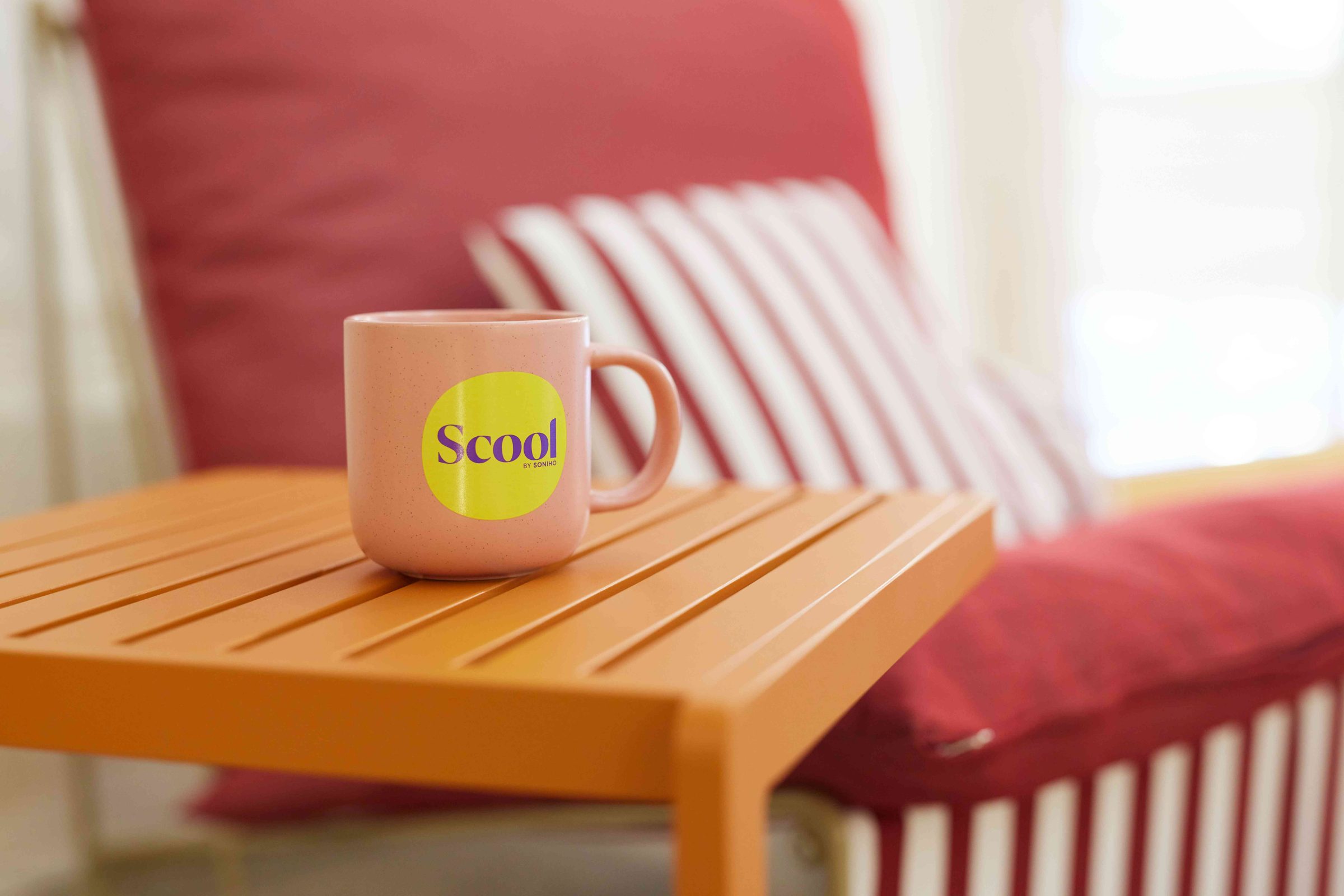 une tasse à café rose poudré avec pour inscription Scool by Soniho en violet sur un fond jaune en forme de rond posée sur une table basse orange et en arrière-plan une assise rouge avec des coussins rayés blancs et rouges