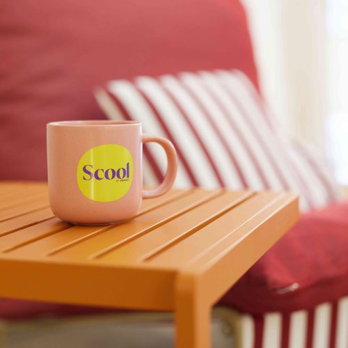 une tasse à café rose poudré avec pour inscription Scool by Soniho en violet sur un fond jaune en forme de rond posée sur une table basse orange et en arrière-plan une assise rouge avec des coussins rayés blancs et rouges
