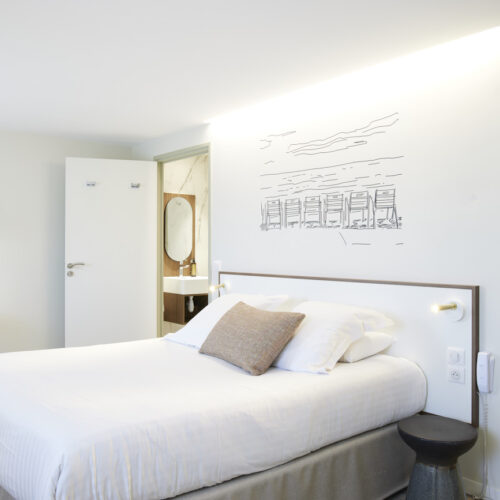 Chambre double dans un hôtel 3 étoiles au cœur de la ville de Nice. La chambre est moderne, un lit double est placé au centre de la photo avec une tête de lit minimaliste. Un dessin filaire de la Promenade des Anglais est dessiné au mur. On remarque également la salle de bain, le dressing et le coin bureau.