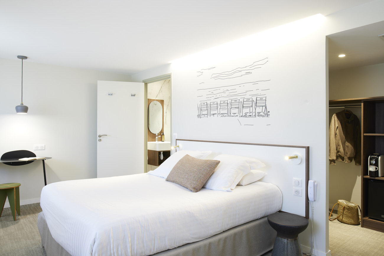 Chambre double dans un hôtel 3 étoiles au cœur de la ville de Nice. La chambre est moderne, un lit double est placé au centre de la photo avec une tête de lit minimaliste. Un dessin filaire de la Promenade des Anglais est dessiné au mur. On remarque également la salle de bain, le dressing et le coin bureau.