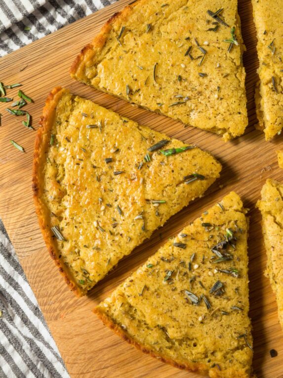 Parts d’une socca, spécialité niçoise avec des herbes aromatisées déposées dessus. La socca est posée sur une planche de bois sur une nappe rayée blanche et grise.
