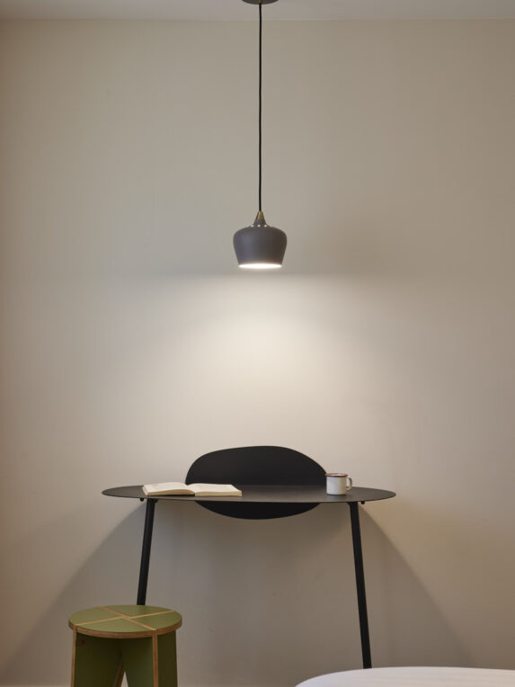 Modern desk with designer black high table, an open book, a cup of coffee. A pendant light hangs over the table, illuminating the desk. A green wooden seat is featured to the left of the pine hotel room desk in Nice.