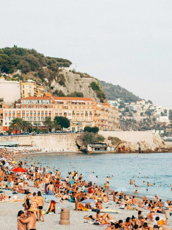 Promenade des Anglais beach with tourists sunbathing and swimming in the Mediterranean Sea. You can see Nice castle and Castel plage.