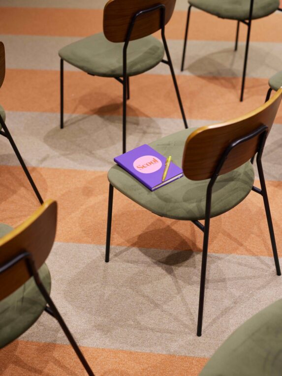 Sedie da sala riunioni dal design retrò con seduta verde e schienale in legno, disposte su un pavimento con motivi geometrici arancioni e beige. Su una sedia ci sono un quaderno viola «Scool» e una penna gialla.