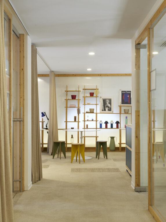 Spazio di lavoro di design con scaffali in legno, sgabelli colorati e tavoli rotondi. La stanza luminosa, con pareti divisorie in vetro e tende beige, crea un’atmosfera accogliente e creativa.