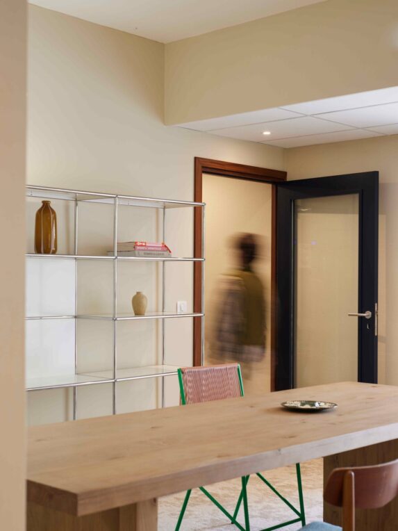Salle avec grande table en bois, étagère métallique minimaliste, deux chaises et silhouette floue près d’une porte ouverte.