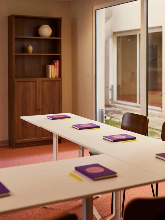 Una sala riunioni luminosa con tavoli bianchi, sedie in legno e taccuini viola disposti davanti a ogni posto a sedere. Una libreria in legno e una grande vetrata che si affaccia su un patio completano la stanza