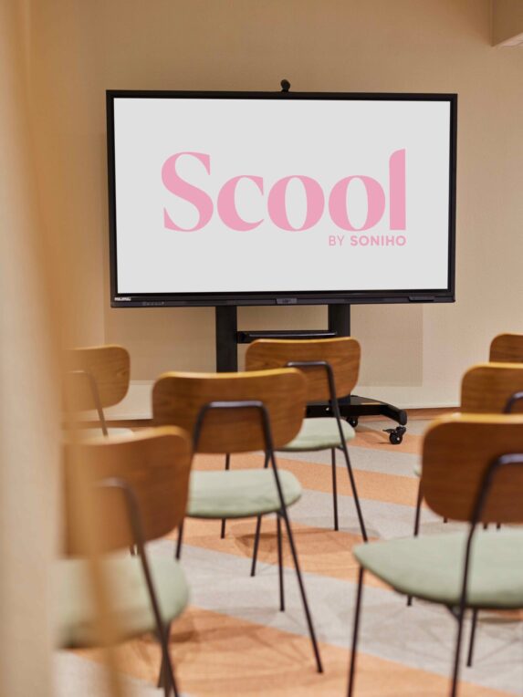 Sala riunioni con sedie in legno e sedute verdi, di fronte a un grande schermo su cui è visibile il logo rosa «Scool by Soniho». L'atmosfera è moderna e minimalista, perfetta per un seminario o una presentazione.