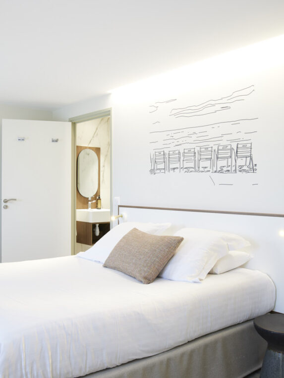 Camera d’albergo moderna e luminosa con un grande letto matrimoniale, una scrivania in stile minimalista, un armadio a vista e un bagno privato. Una illustrazione murale raffigurante delle cabine da spiaggia decora la parete sopra il letto.