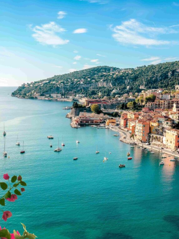 Vista panoramica della costa mediterranea con il mare turchese, barche a vela all’ancora e case colorate lungo la costa, sotto un cielo azzurro.