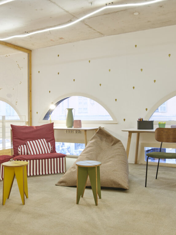Un’area relax colorata con poltrone rosse a righe, un pouf beige, sedie di design e tavolini gialli e verdi. La stanza luminosa ha finestre arrotondate e un arredamento moderno e giocoso.