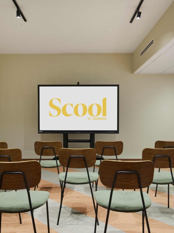 Una moderna sala conferenze con sedie in legno e sedute verdi, di fronte a un grande schermo su cui compare il logo giallo «Scool by Soniho». La stanza è sobria e luminosa, con un pavimento dai motivi geometrici arancioni e grigi