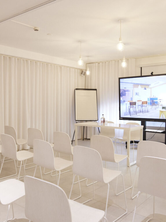 Una sala conferenze luminosa e dalle linee essenziali, con file di sedie bianche, un maxischermo per le presentazioni, una lavagna a fogli mobili e un tavolo per il relatore. L’atmosfera è moderna e minimalista