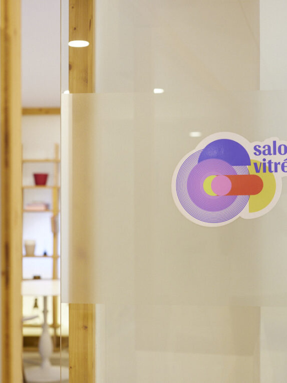 Porta a vetri con un adesivo colorato che dice “Salone con vetrate”, che si affaccia su uno spazio interno luminoso con mensole in legno e oggetti decorativi