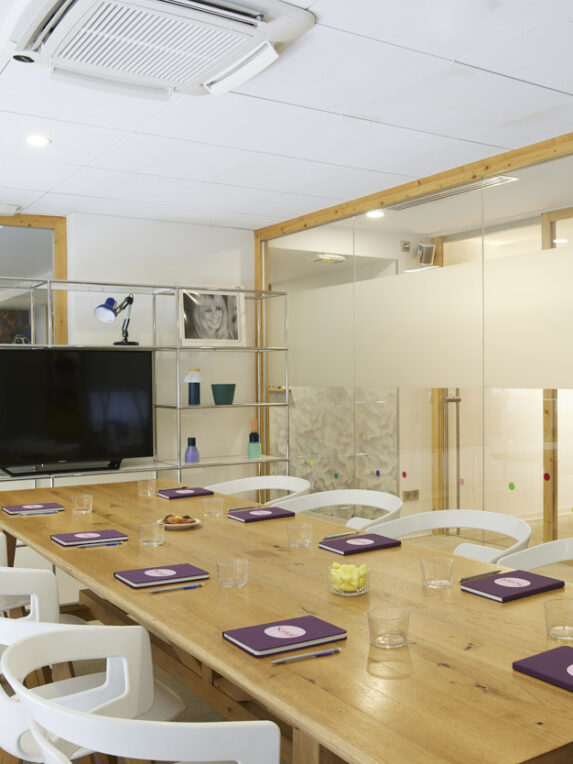 Sala riunioni con pareti vetrate, un grande tavolo in legno, sedie bianche e taccuini viola “Scool” posizionati davanti a ogni posto. Un televisore, scaffali metallici e una macchina del caffè completano l’arredamento moderno e accogliente