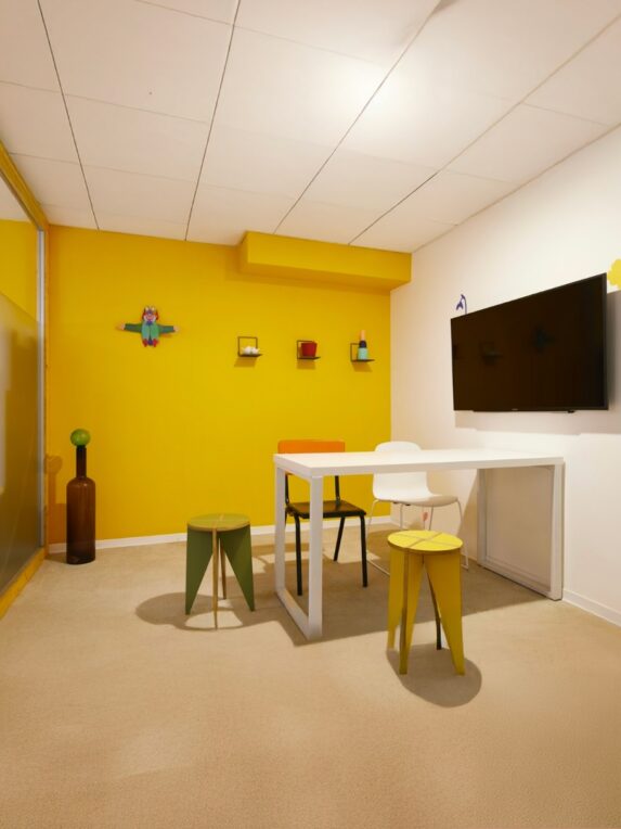 The Small Lounge at the Hôtel du Pin: a small, empty meeting room with a yellow wall, a white table and chairs, three colorful stools, and a TV screen mounted on the wall.