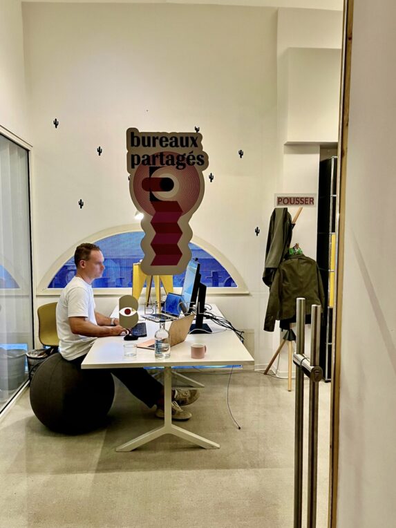A man working on a computer in a shared office, seen through a glass door labeled “shared offices”; a half-moon window and a coat rack are visible nearby.