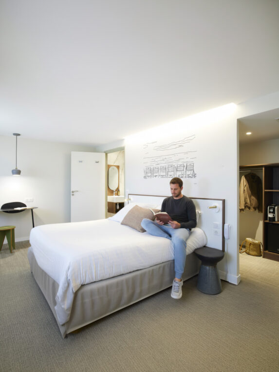 A man is sitting on a bed in a modern hotel room, reading; we can see a desk area, an open bathroom, and a walk-in closet to the side.