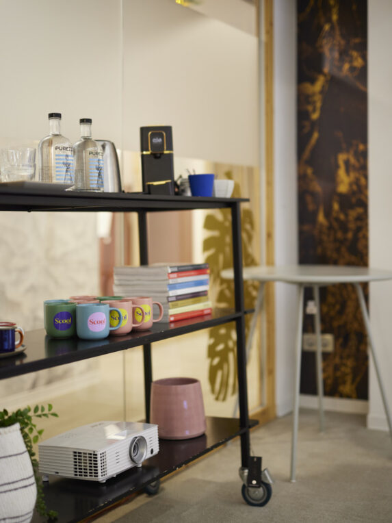 A refreshment cart in a meeting room, featuring bottles of water, a coffee machine, colorful “Scool” cups, a stack of books, and a projector.
