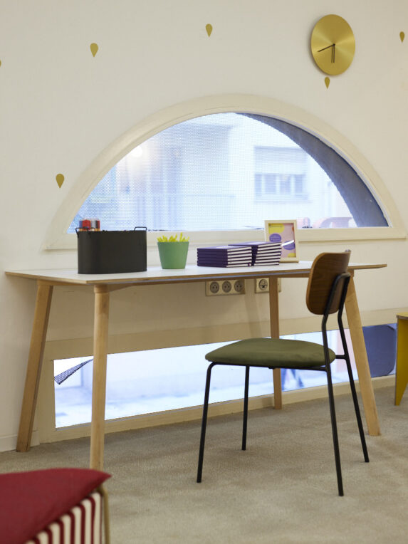 A small workspace with a wooden table and a chair, set in front of a half-moon window; on the table are notebooks and supplies, with a gold clock on the wall.