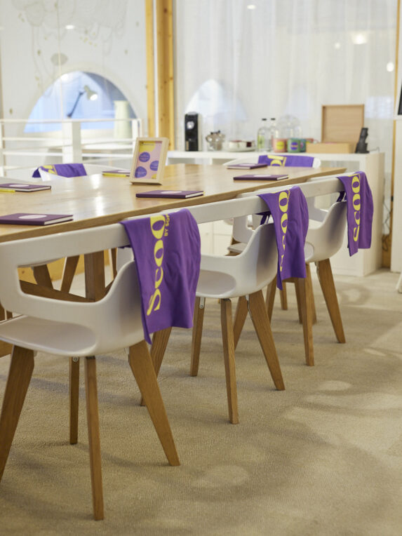 A large conference table with white chairs, notepads placed at each seat, and purple “Scool” tote bags hanging from the backs of the chairs; a kitchenette at the back of the glass-walled lounge.