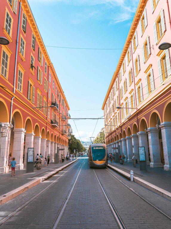A tram traveling down the center of the wide Avenue Masséna, lined with colorful buildings featuring arcades, under a blue sky.