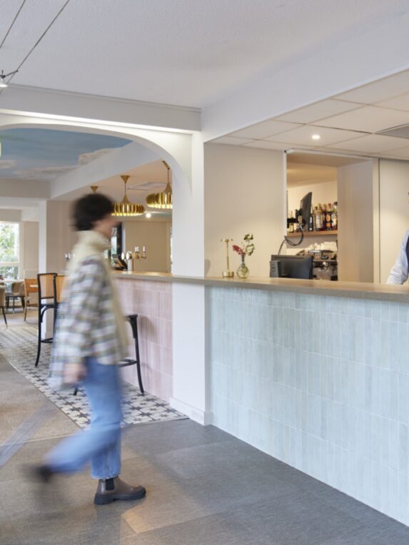 Réception d’hôtel moderne avec comptoir carrelé, un employé derrière le bar et une personne passant devant, dans un espace lumineux avec coin bar et tables en arrière-plan.