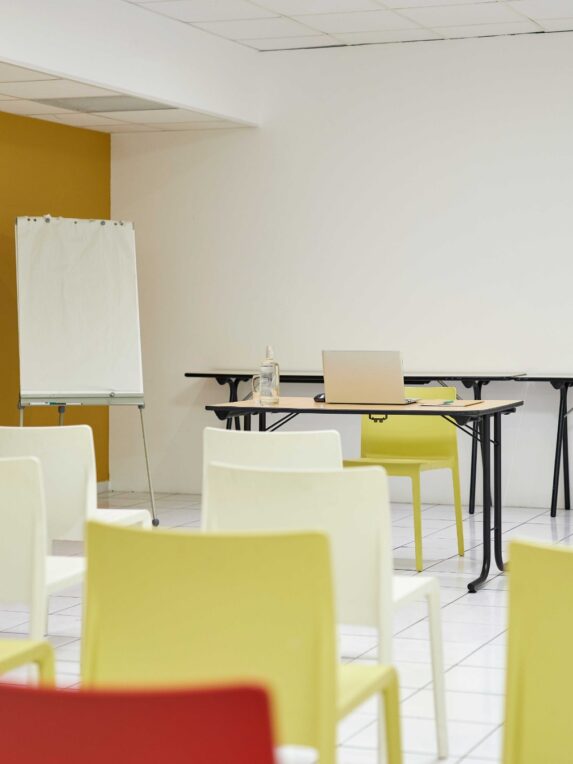 Salle de formation avec chaises alignées face à une table équipée d’un ordinateur portable, un paperboard et un écran de projection.
