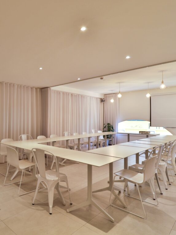 Salle de réunion lumineuse avec tables disposées en U, entourées de chaises blanches, et un écran de projection au fond de la pièce.