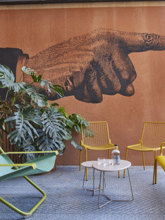 Espace terrasse lounge avec chaises jaunes et table basse, devant un mur décoré d’une grande illustration d’une main pointant, avec des plantes vertes à gauche.