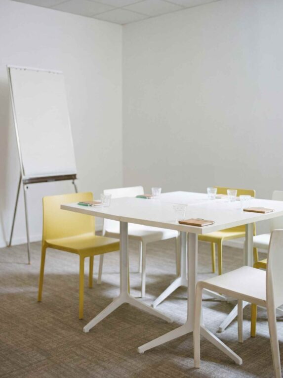 Petite salle de réunion lumineuse avec table blanche entourée de chaises jaunes et blanches, verres d’eau et carnets disposés sur la table, et un tableau de présentation sur chevalet à gauche.