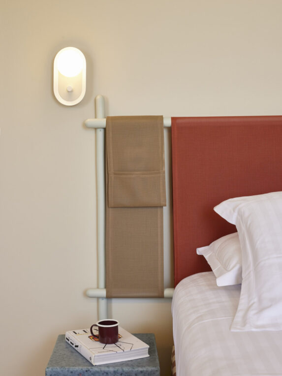 A close-up of a bedroom featuring a red headboard, a lit wall sconce, white pillows, a blue nightstand, and a mug resting on a book.