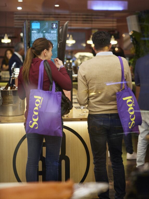 Two people seen from behind in a bustling bar, carrying purple Scool tote bags, standing in front of a counter with screens and glasses.
