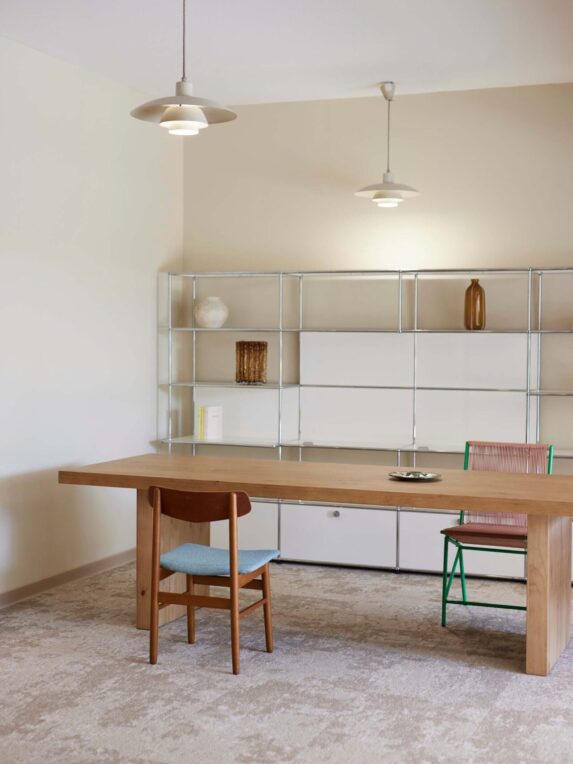 A bright room with a large wooden table, two mismatched chairs, a minimalist wall shelf, and two white pendant lights.