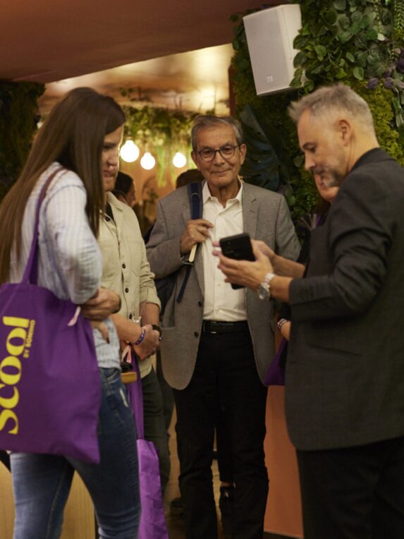 A group of people chatting inside the Foam space at the Hôtel du Pin, beneath the Scool workspaces; some are carrying purple bags with the word “Scool” on them, while a man in the foreground is looking at his phone.