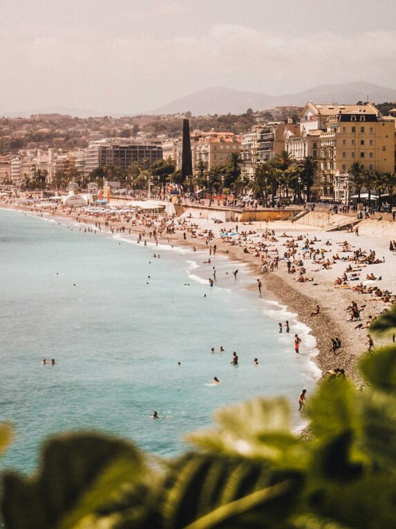 Plage animée bordant une ville côtière, avec de nombreuses personnes se baignant et se reposant au soleil, des palmiers et des immeubles le long de la promenade, et des montagnes en arrière-plan.