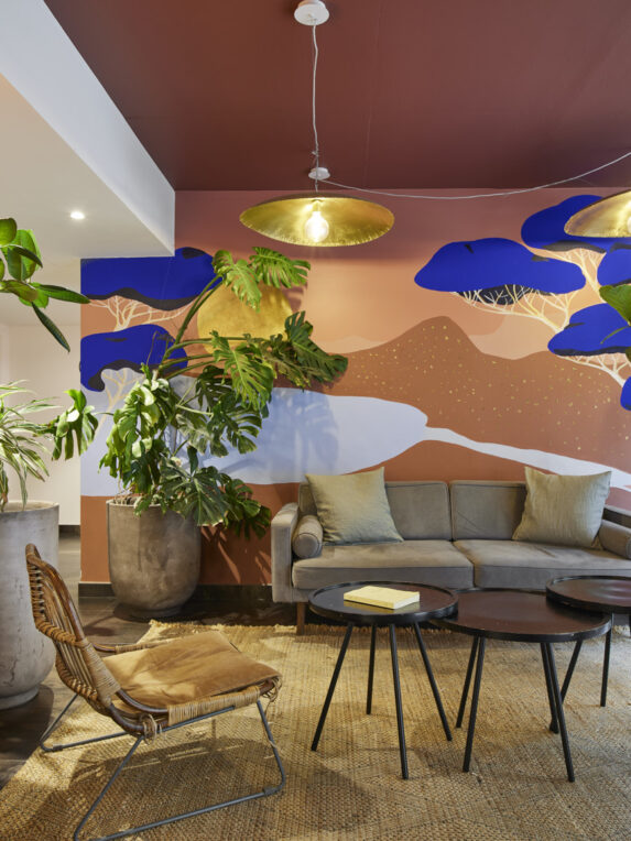 Salon chaleureux avec canapé gris, tables basses noires, fauteuil en rotin et grandes plantes, devant un mur décoré d’une fresque colorée et éclairé par des suspensions dorées.