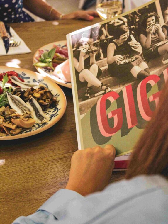 Personne tenant un menu illustré à table, avec plats de nourriture et verres autour, dans un cadre de repas convivial.