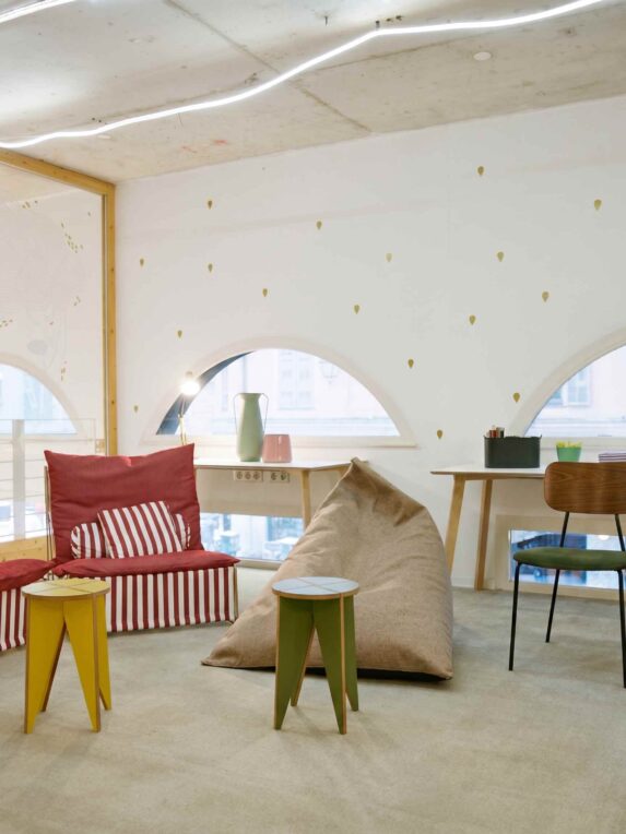 Espace de coworking lumineux avec mobilier coloré, comprenant canapé, pouf, petites tables et coin bureau près de fenêtres arrondies avec rideaux.