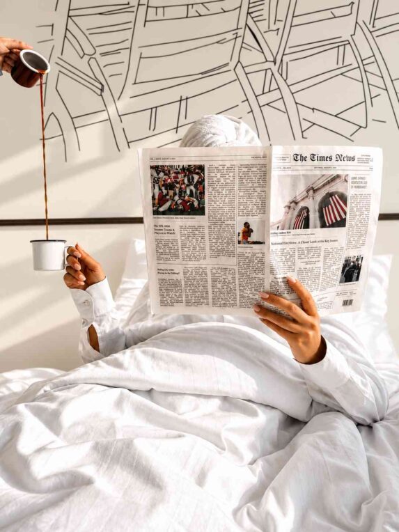 Personne lisant le journal au lit avec une tasse de café, dans une chambre lumineuse et moderne.