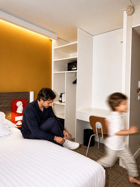 Père assis sur le lit d’une chambre d’hôtel pendant qu’un enfant passe devant lui, dans un espace moderne et lumineux.