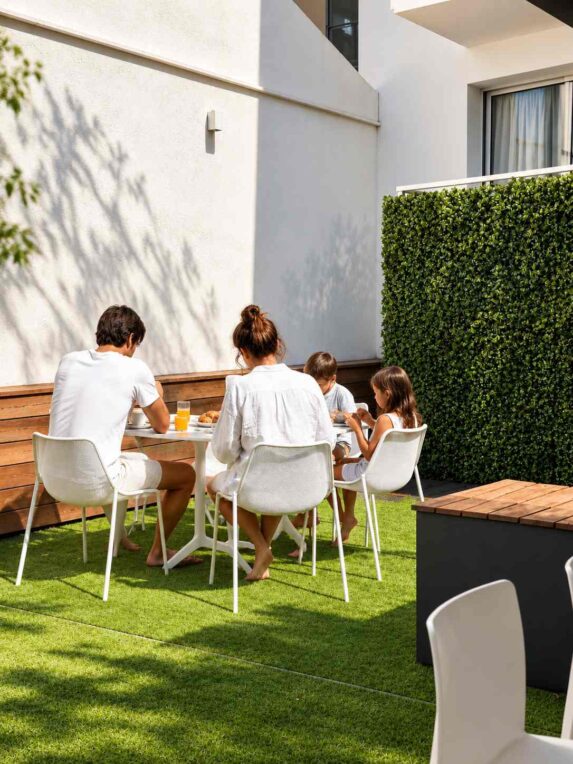 Famille de quatre personnes prenant le petit-déjeuner autour d’une table sur une terrasse extérieure ensoleillée, dans une ambiance calme et conviviale.