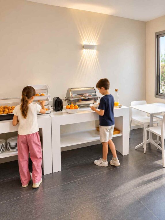 Deux enfants se servent au buffet du petit-déjeuner dans la salle lumineuse et moderne de l’Hôtel Abrial.