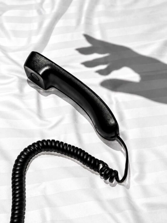 Combiné de téléphone noir posé sur un lit blanc, éclairé par la lumière naturelle avec l’ombre d’une main en arrière-plan.